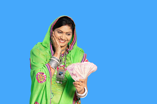 Happy excited indian woman wearing sari holding indian rupees note against blue background, first salary, housewife with cash, female with lots of money, savings and financial growth.
