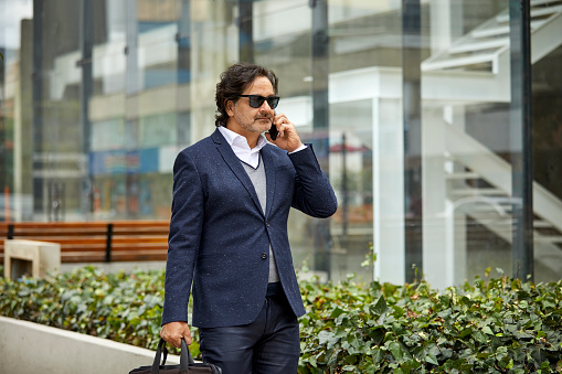 Mature businessman talking on mobile phone while walking. Male professional is outside office building. He is wearing suit.