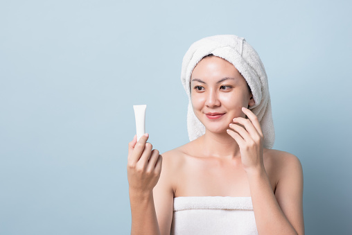 Beautiful Asian young woman with bath towel on her head smiling holding white cream in her hand, touching her glowing skin face with copy space light blue pastel background.