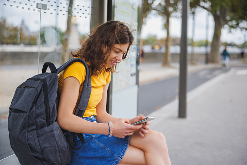 Female university student smiling and using phone while waiting for the bus to campus in the street in Paris, France