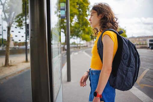 Beautiful female university student with backpack standing in front of media banner and reading routes on a bus stop in the street in Paris, France