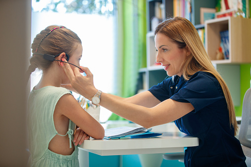 Female speech therapist working with girl at the appointment, adjusting her headset before reading, speech therapy techniques and technology