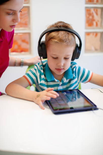 istock Preschool boy wearing headphones and using tablet, guided by speech therapist 1643816354