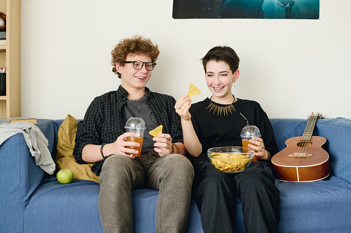 Cheerful teenage couple in casualwear having potato chips with soda while sitting on blue soft couch at home and chatting about something funny