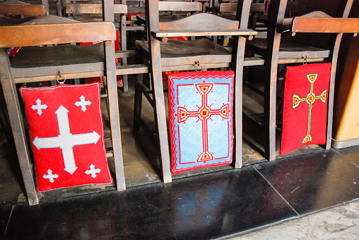 Color image depicting a senior man in his 60s sitting alone in a church while wearing a protective face mask and face shield. The pews are mark with ticks and crosses to delineate a social distancing system during the covid-19 pandemic. Room for copy space.