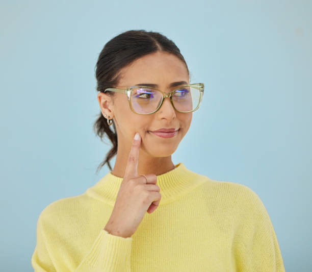 istock Woman, smile and glasses in studio thinking, problem solving style and vision on blue background. Ideas, eyewear and happy model brainstorming with hand on face in cool fashion, mindfulness and gen z 1643871641
