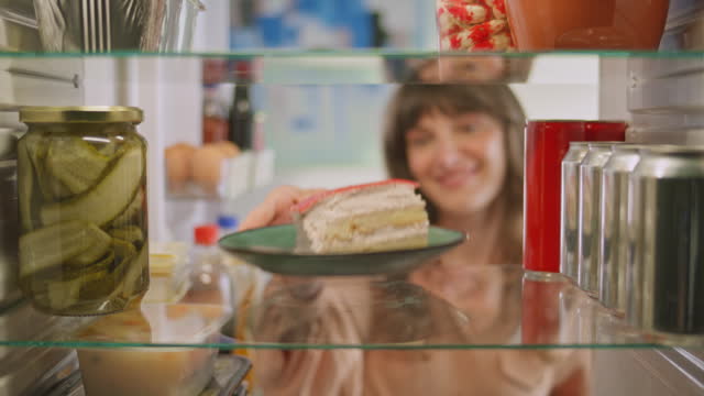 Woman checking the fridge and taking a plate with a slice of cake from the shelf