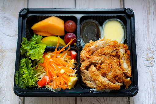 Picture of a lunch box menu featuring fried chicken with fresh vegetables and fruits on the table.