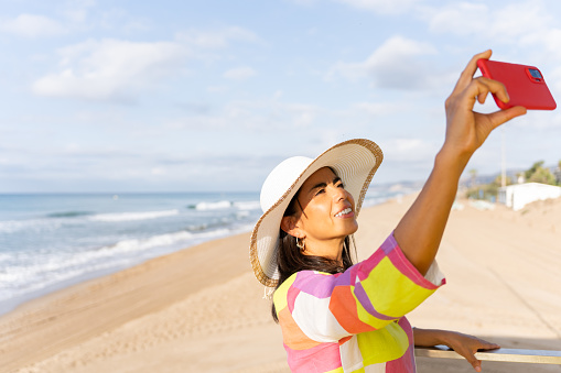 Beautiful latina woman taking a portrait from the top of the lifeguard watchtower wearing a flashy colorful dress and hat on a lovely summer day by the beach