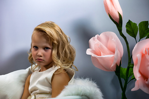 Pensive lovely little girl with hairstyle sitting on chair, thought looking at camera. Cute child lady bored in grey room with flowers, studio shot, gen a. Youth emotion concept. Copy ad text space