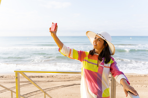 Beautiful latina woman with a hat and flashy and colorful dress enjoys a beautiful summer guide from the top of a lifeguard watchtower doing a selfie.