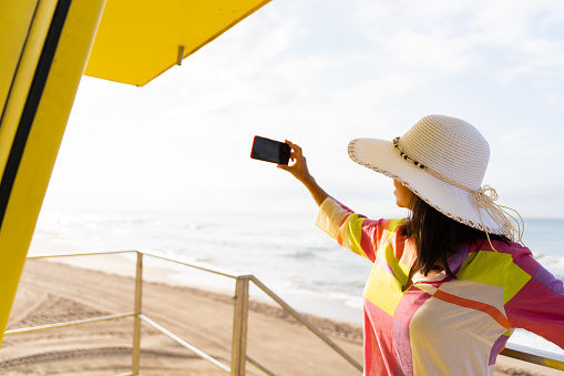 Unrecognizable woman takes a selfie from the bright yellow lifeguard tower on a beautiful summer day in colorful dress and hat.