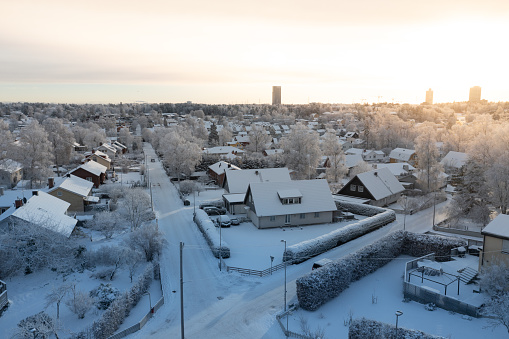 As the first rays of sunrise touch the horizon, a residential district awakens to a pristine winter scene. Freshly fallen snow blankets the houses and streets, capturing the untouched beauty of the season. The golden light brings a warmth to the cold surroundings, painting a tranquil and picturesque tableau of urban life amidst nature's embrace.