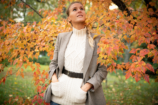 A beautiful young stylish woman walks in the park among the trees on an autumn sunny day and smiles holding her hands in her pockets. The blonde girl is wearing a gingham jacket with gold buttons, a warm white sweater and denim shorts.