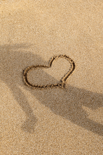 Discover love at the shoreline with 'Heart in Sardinian Sands.' This portrait-oriented photo captures the fleeting beauty of a hand-drawn heart in wet Sardinian sand. Freshly touched by retreating waves, the sand serves as a canvas for this intimate moment, with the shadow of the artist subtly captured.