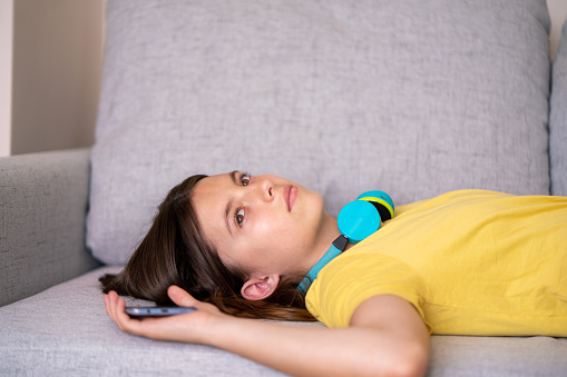Teenage girl whit headphones and mobile phone lying on the sofa at home and looking away