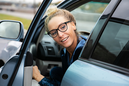 Beautiful blue-eyed blonde in black-rimmed glasses with diopters in a blue jacket. The woman's hair is gathered in a bun at the top. The beauty smiles pleasantly and emotionally, she is in a good mood. Businesswoman parked in her blue car during the day and gets out of it. She looks back over her shoulder as she opens the driver's door.