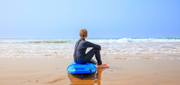 Young boy sitting on surfboard looking at the sea- water sport,  vacation,  family surfer holiday concept
