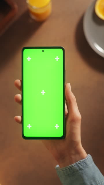 Vertical Screen: Top static View of Anonymous Woman Using Smartphone with Mock Up Green Screen Chromakey Display on Kitchen Table With Food Ingredients. Female Watching a Cooking Recipe Video Tutorial