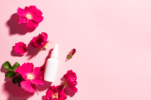 White cosmetic serum bottle with dropper, wild rose flowers on pink background, copy space. Natural organic cosmetics, beauty herbal skin care product, spa aroma oil.