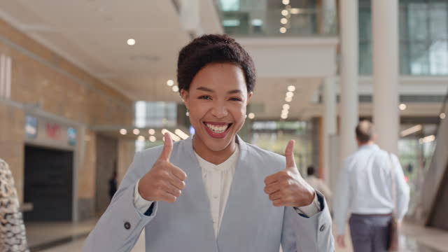 Business woman, face and thumbs up for success, like and thank you or vote, yes and excellence sign in office building. African person walking in corporate lobby with yes, agreement or support emoji