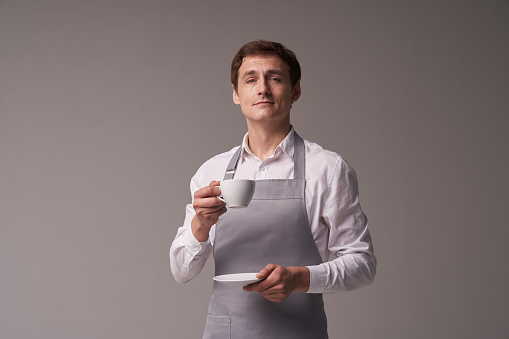 The positive male waiter on a grey background. The barista holds a coffee drink for the customer. Portrait of a successful young man. Layout with copy space. High quality photo