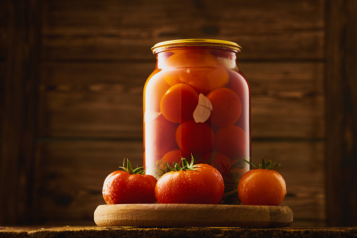 Glass jar of homemade pickled tomatoes and fresh tomatoes. Wooden rustic backdrop. Studio shot from a low angle.