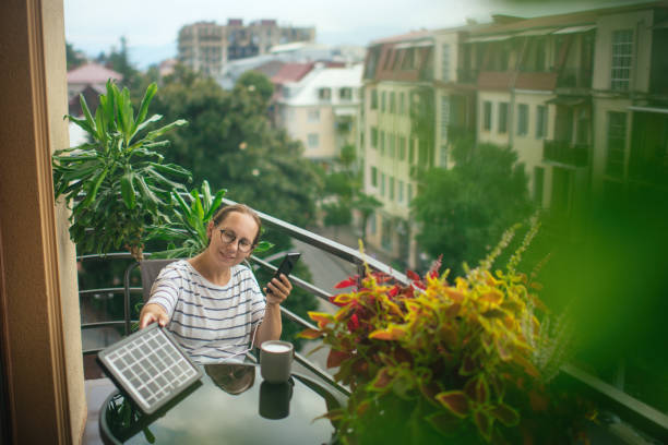 Woman charging smartphone with solar panel on a balcony in a sunny summer day. Sustainable lifestyle, solar energy, ecological alternative energy and ecology concepts. stock photo