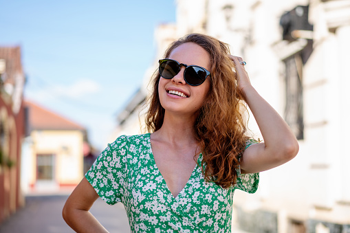 Young curly hair woman wearing green summer dress and sunglasses posing in front of town street.
