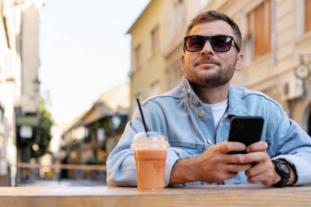istock Man wearing denim jacket and sunglasses online messaging using his mobile phone while sitting in open air cafe in summer. 1644085442