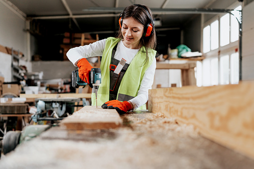 A young Caucasian female carpenter is smiling while using a drill on a wooden board. She's in her workshop.