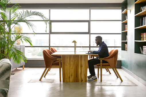 Full length side view of bearded Black man in casual attire sitting at table, concentrating on typing, floor to ceiling windows with view of downtown Nairobi.