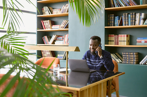 Waist-up view of bearded Black man in casual attire sitting at table in coworking office, looking at computer screen and smiling while listening to caller.