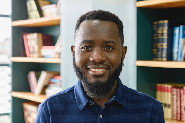Headshot of bearded Nairobi businessman Close view of mid adult man wearing blue open collar shirt, smiling at camera, coworking office bookshelves in background. kenyan man stock pictures, royalty-free photos & images