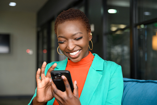Waist-up view of shorthaired Black woman wearing hoop earrings, teal jacket over orange top, and smiling while scrolling portable device in modern office.