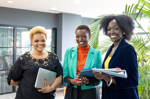 Waist-up view of professional associates in 20s, 30s, and 40s standing together in modern office and smiling at camera.