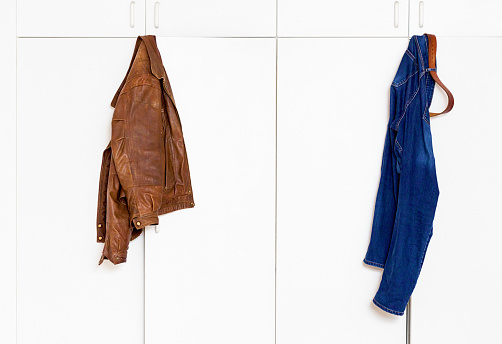 Distressed leather jacket and denim jeans hanging up on the tops of the doors of a white closet, with copy space.