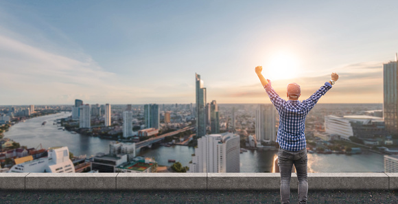 Rear view of male traveler with raised hands standing on the top of a tower and looking at the urban city with sunset sky. Woman happy enjoying the city view from top of a city.