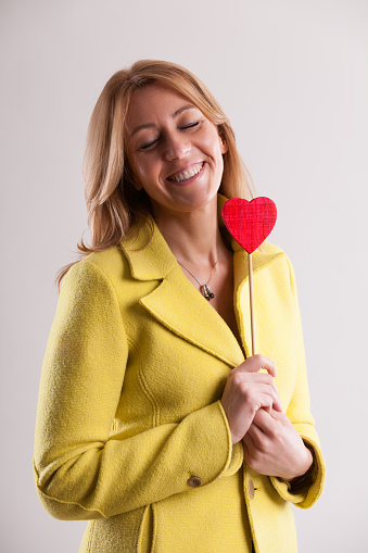 smiling, dreamy adult woman in a yellow coat tightly holds a red heart symbol, expressing deep love, warmth, embrace, and mutual understanding. Finding such connection is rare in relationships. Also,