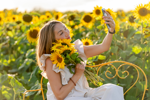 cheerful  asian woman sitting on bench in a white dress   with a bouquet and take selfie in the field of sunflowers in Yorkshire