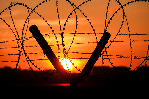 Barbed wire and razor wire of the airport boundary at Gate 13 of Sydney Kingsford-Smith Airport.  A band of red is caused by the sun reflecting off the airport runway.  This image was taken from near Shep's Mound, Ross Smith Avenue, Mascot facing the setting sun at sunset on 25 August 2023.
