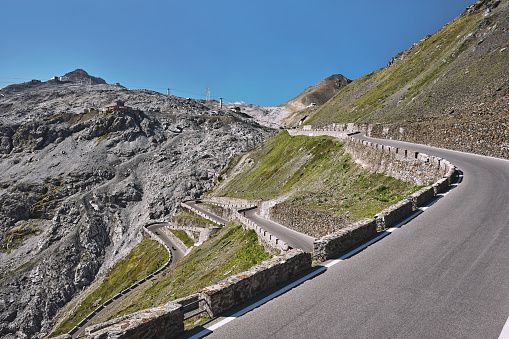 Wide-angle view of the renowned road ascending Passo dello Stelvio (formally known as Strada Statale 38 dello Stelvio), a mountain pass which connects Valtellina and Val Venosta at an elevation of 2,757 metres above sea level, in the Italian Alps, offering breathtaking views on the surrounding mountaintops and the underlying valley. The dazzling bright light of a summer noon, a perfectly clear sky, rough rocky cliffs and steep green slopes, cyclists, bikers and cars following the narrow, winding strip of asphalt. High level of detail, natural rendition, realistic feel. Developed from RAW.