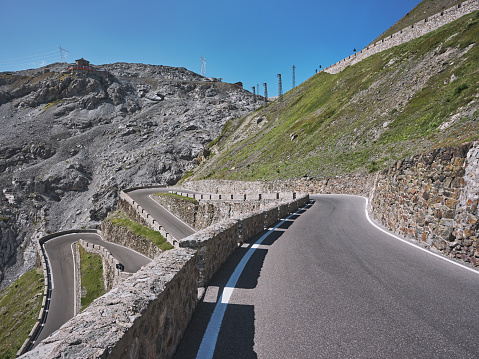 Wide-angle view of the renowned road ascending Passo dello Stelvio (formally known as Strada Statale 38 dello Stelvio), a mountain pass which connects Valtellina and Val Venosta at an elevation of 2,757 metres above sea level, in the Italian Alps, offering breathtaking views on the surrounding mountaintops and the underlying valley. The dazzling bright light of a summer noon, a perfectly clear sky, rough rocky cliffs and steep green slopes, a narrow, winding strip of asphalt. High level of detail, natural rendition, realistic feel. Developed from RAW.