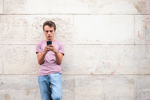 Portrait of handsome young man leaning to wall outside and looking at mobile phone