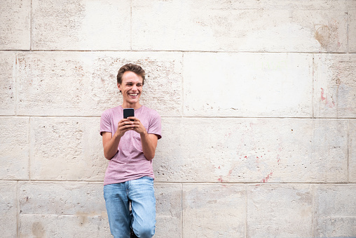 Portrait of smiling young man leaning against wall with cellphone outside