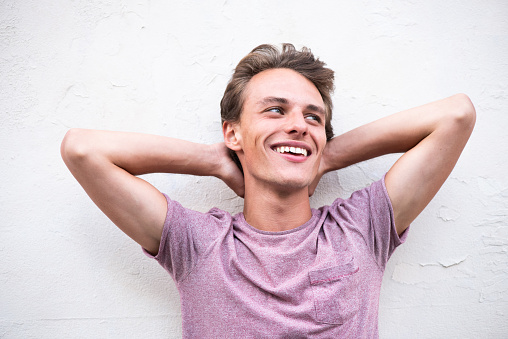 Close up portrait of happy young man with hands behind head leaning to white wall