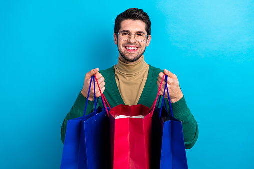 Photo of wearing green jumper young guy showing his purchases open packages bargains packages shopaholic isolated on blue color background.