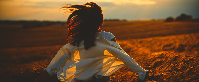 Carefree young woman running through wheat crops exploring farm and enjoying vacation during sunset