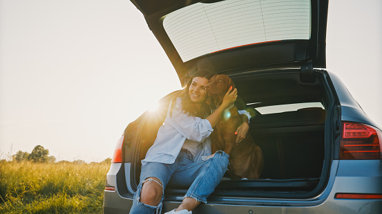 Cheerful young woman embracing brown Vizsla dog while sitting in trunk of car at agricultural field during vacation