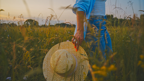 Midsection of young woman holding straw hat walking through wheat crops in field at sunset during vacation